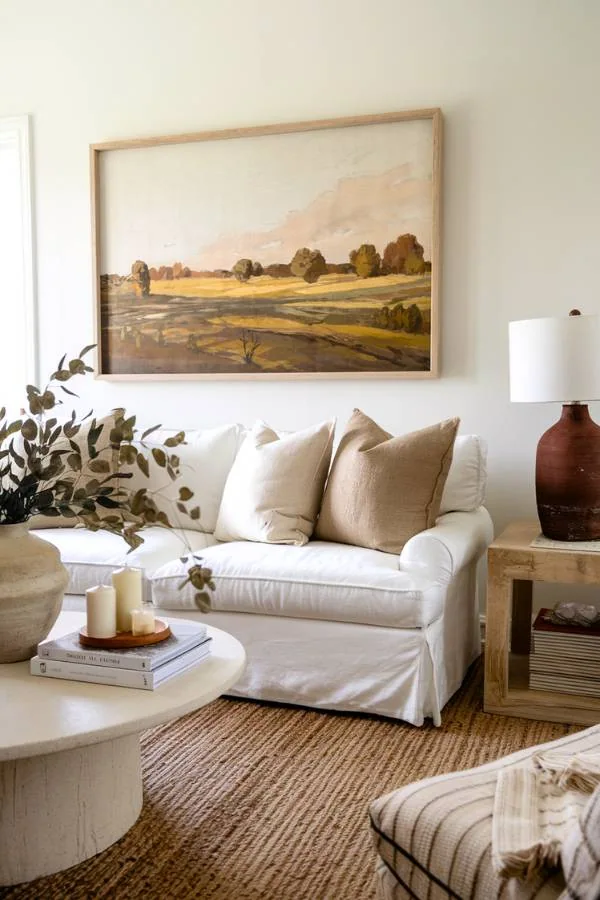 Organic modern living room idea featuring a white slipcovered sofa, landscape artwork, natural jute rug, round concrete coffee table, and eucalyptus branches in earthy neutral tones
