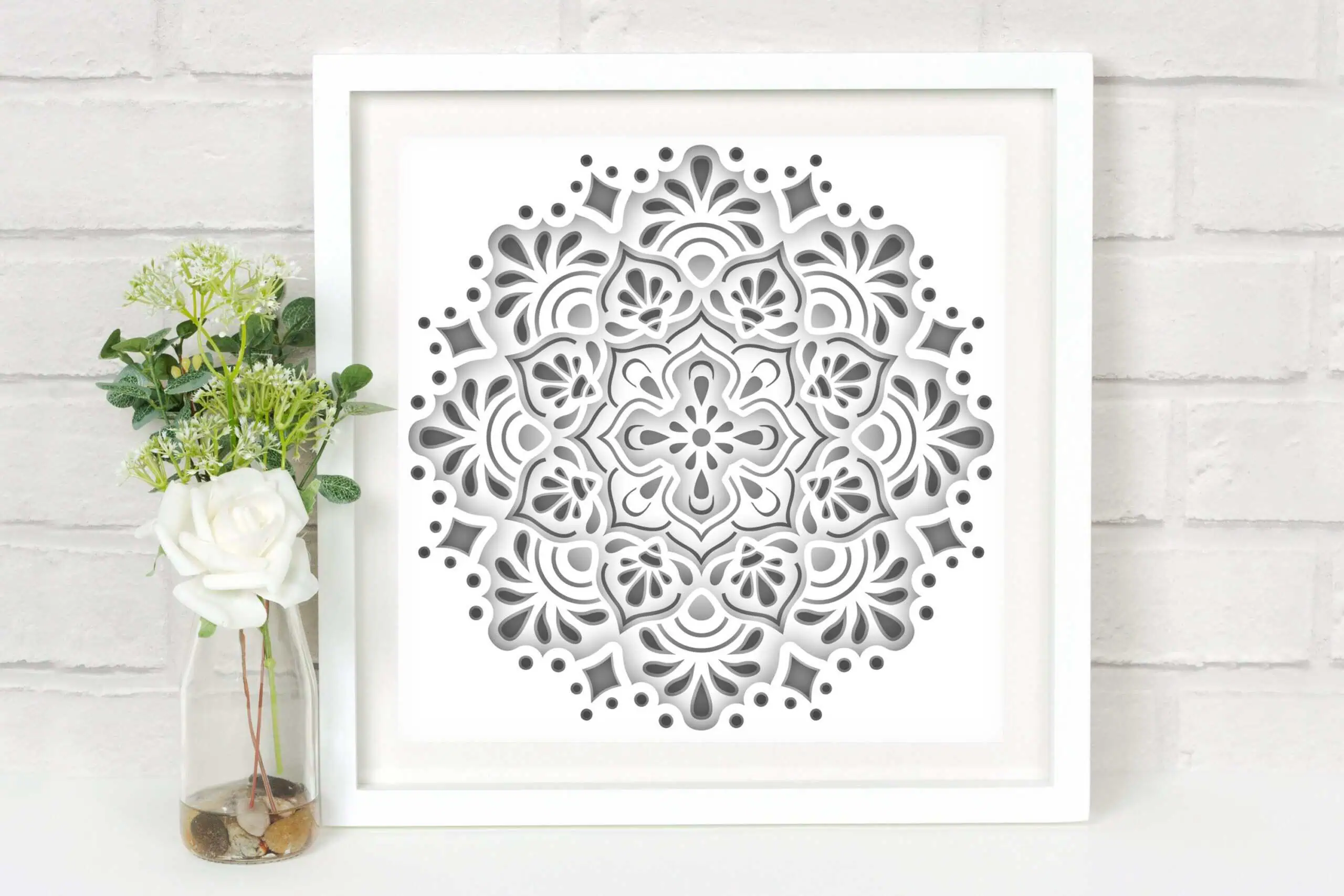 white and gray layered mandala shadow box in a white frame next to white flowers