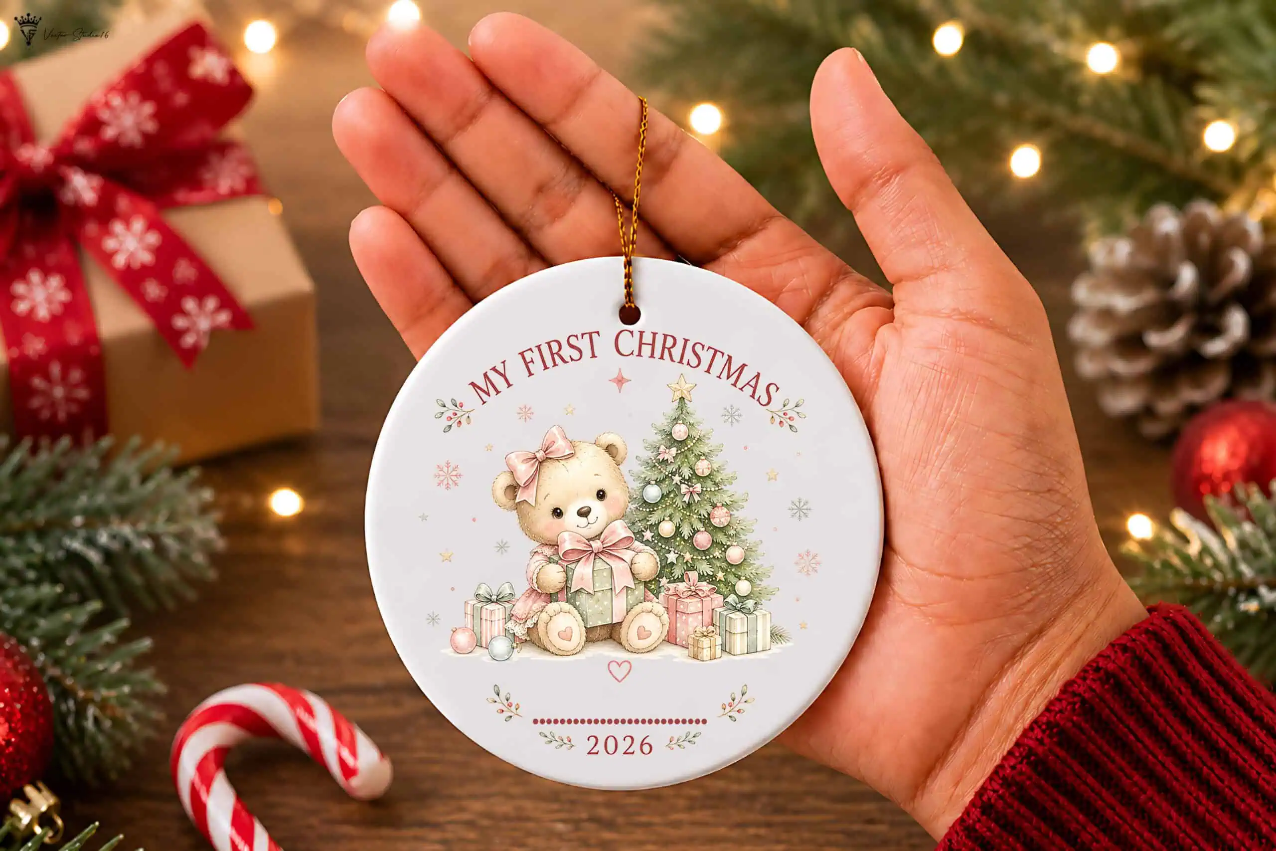 hand holding a white round ornament with a teddy bear in a pink bow beside a Christmas tree and presents, with my first Christmas 2026